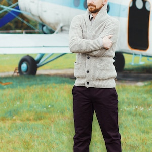 May include: A man wearing a light gray cardigan sweater with a collared shirt underneath, dark pants, and brown shoes. He is standing in front of a small airplane.