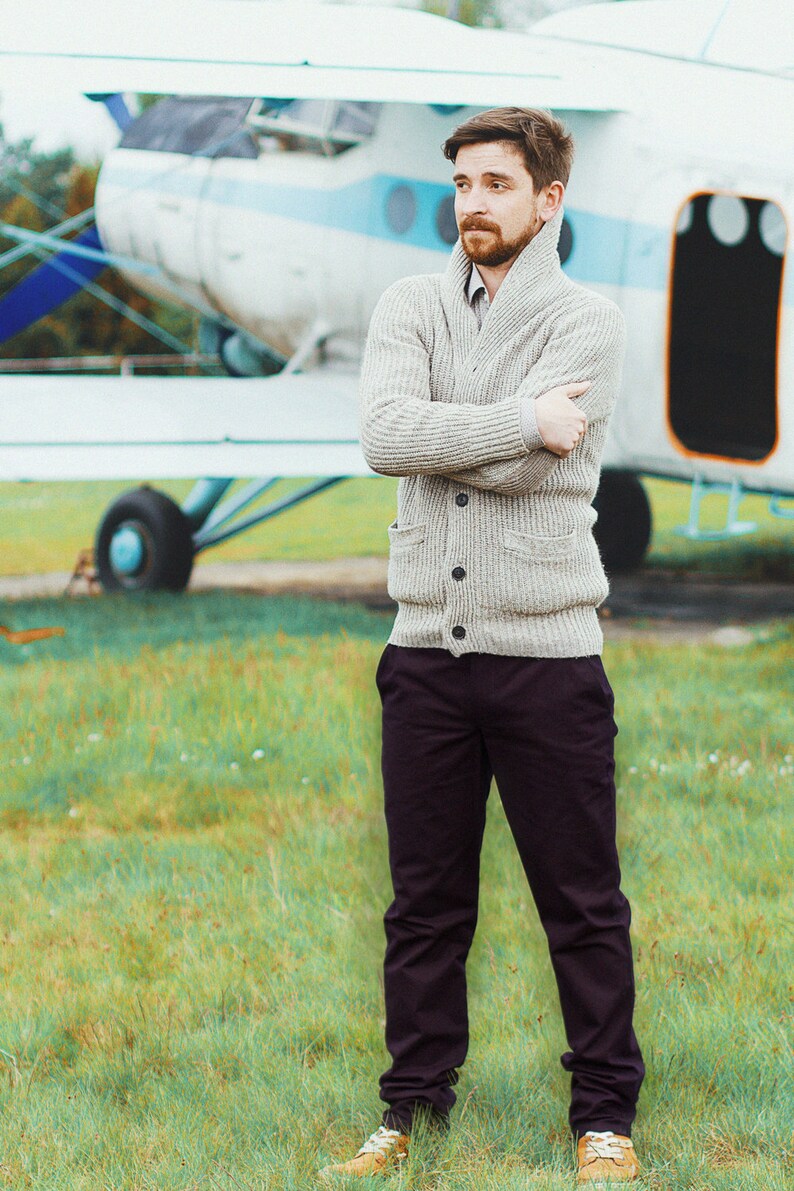 May include: A man wearing a light gray cardigan sweater with a collared shirt underneath, dark pants, and brown shoes. He is standing in front of a small airplane.