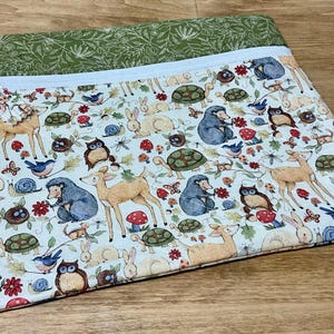 May include: A white zippered pouch with a green and white fabric lining. The pouch is decorated with a repeating pattern of woodland animals, including owls, deer, squirrels, and birds. The pouch is perfect for storing small items like makeup, jewelry, or craft supplies.