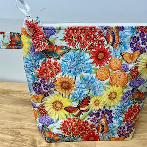 May include: A floral print bag with a white zipper and a matching handle. The bag features a colorful pattern of butterflies and flowers in shades of red, orange, yellow, blue, and purple. A decorative butterfly charm is attached to the zipper.