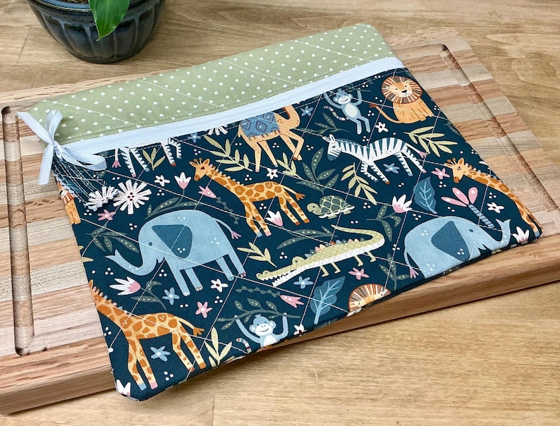 May include: A quilted fabric pouch with a zip closure. The pouch features a dark teal background with a repeating pattern of safari animals, including giraffes, elephants, and zebras. The top edge is a light green with white polka dots.