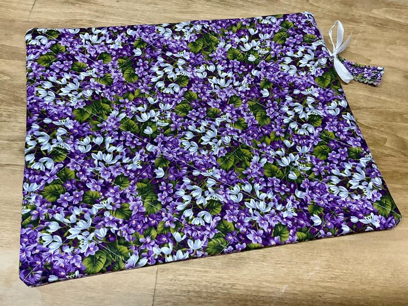 Puede incluir: Una tela de estampado floral morado y blanco con un dise&ntilde;o acolchado. La tela tiene una cinta blanca unida a una esquina.