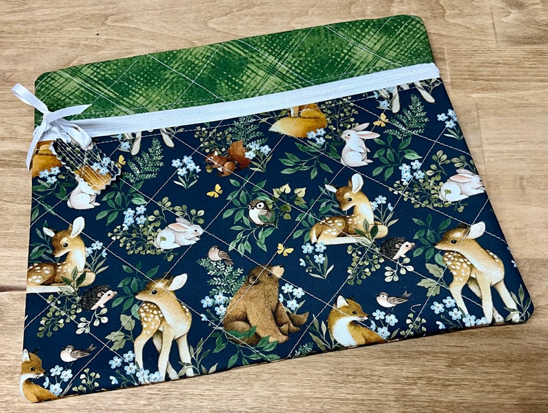 May include: A quilted fabric pouch featuring a woodland animal print. The design includes deer, rabbits, foxes, and birds amidst foliage and flowers on a navy blue background. The top edge has a green plaid pattern and a white zipper.