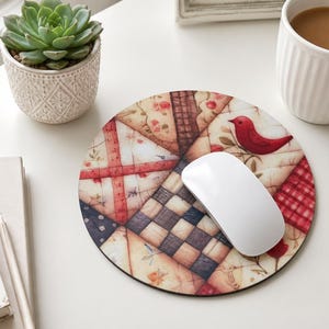 May include: A round mouse pad featuring a patchwork quilt design with cream, red, and dark blue fabric patterns, including a small red bird illustration. A smooth white computer mouse rests on top of the circular pad, which displays a mix of checkered, floral, and striped textile-inspired graphics.