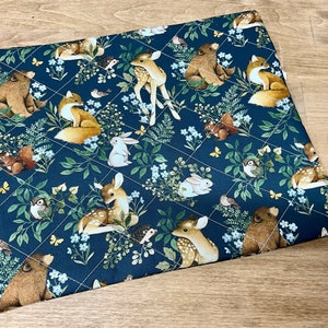 May include: A quilted, square fabric featuring a dark blue background with a repeating pattern of woodland animals and floral elements. The design includes deer, foxes, bears, rabbits, and birds, with white stitching creating a diamond grid.