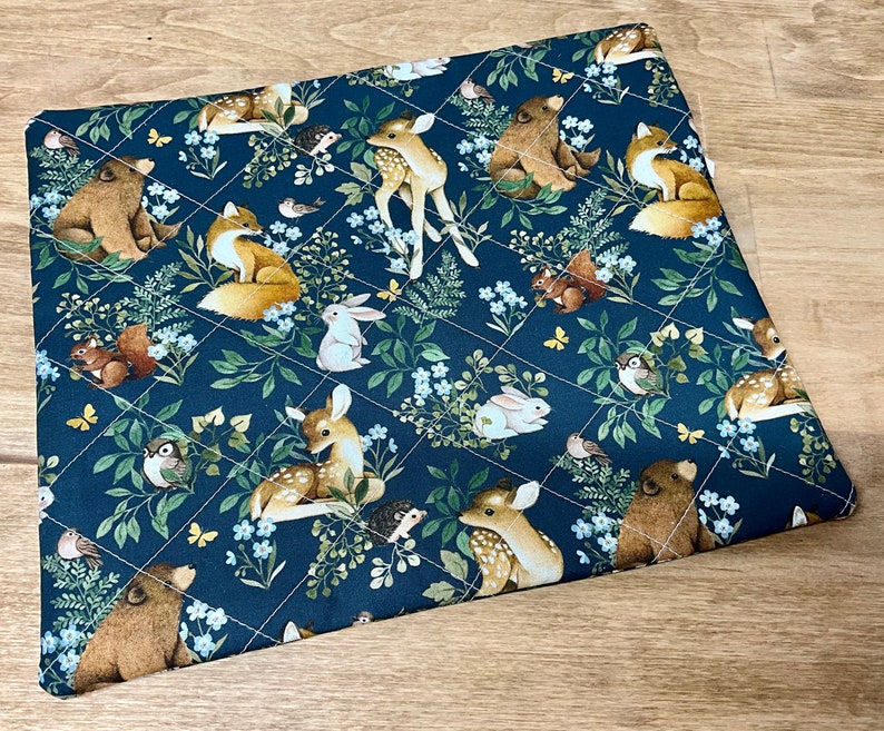 May include: A quilted, square fabric featuring a dark blue background with a repeating pattern of woodland animals and floral elements. The design includes deer, foxes, bears, rabbits, and birds, with white stitching creating a diamond grid.