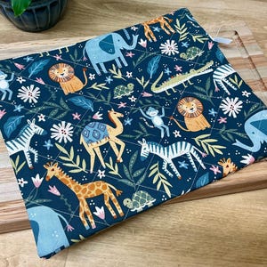 May include: A square, quilted potholder with a dark teal background. The fabric features a repeating pattern of cartoon animals, including lions, giraffes, zebras, elephants, and monkeys, along with floral and leaf accents. A white loop is attached to one corner.