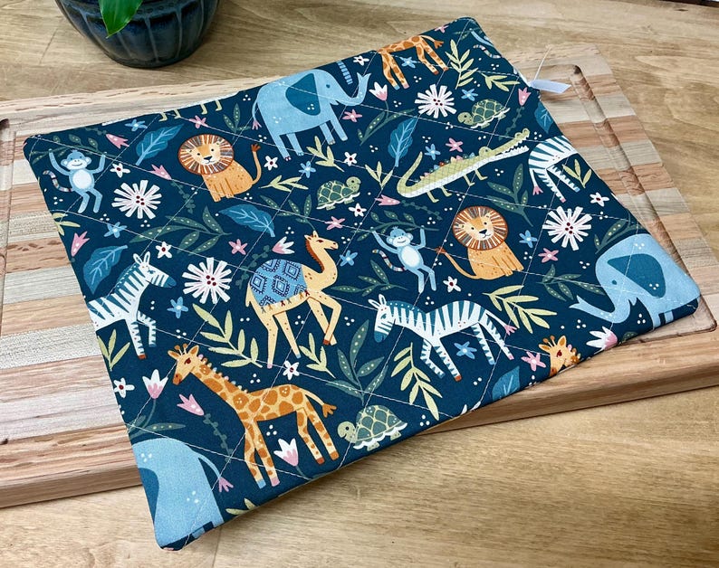 May include: A square, quilted potholder with a dark teal background. The fabric features a repeating pattern of cartoon animals, including lions, giraffes, zebras, elephants, and monkeys, along with floral and leaf accents. A white loop is attached to one corner.