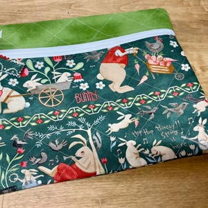 May include: A green zippered pouch with a quilted fabric exterior. The pouch features a whimsical spring-themed print with bunnies, birds, flowers, and the text "Hooray!! Spring is here".