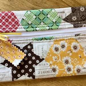 May include: A patchwork fabric zippered pouch with a yellow ribbon and tag. The pouch is made of various fabrics with floral, geometric, and plaid patterns in shades of red, yellow, green, blue, and brown.