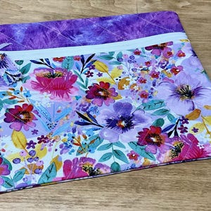 May include: A purple and white quilted zippered pouch with a floral print featuring pink, purple, and yellow flowers. The pouch has a white tassel.