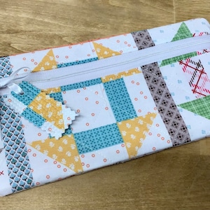 May include: A white zippered pouch with a patchwork design featuring various patterns in shades of blue, yellow, green, and red. The pouch has a white ribbon tied around it and a small fabric tag attached.