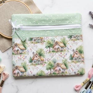 May include: A square quilted zipper pouch featuring a light green fabric top with white polka dots and a main body printed with a pattern of small cottages, trees, and flowers. The pouch includes a white zipper closure with a small fabric tag.