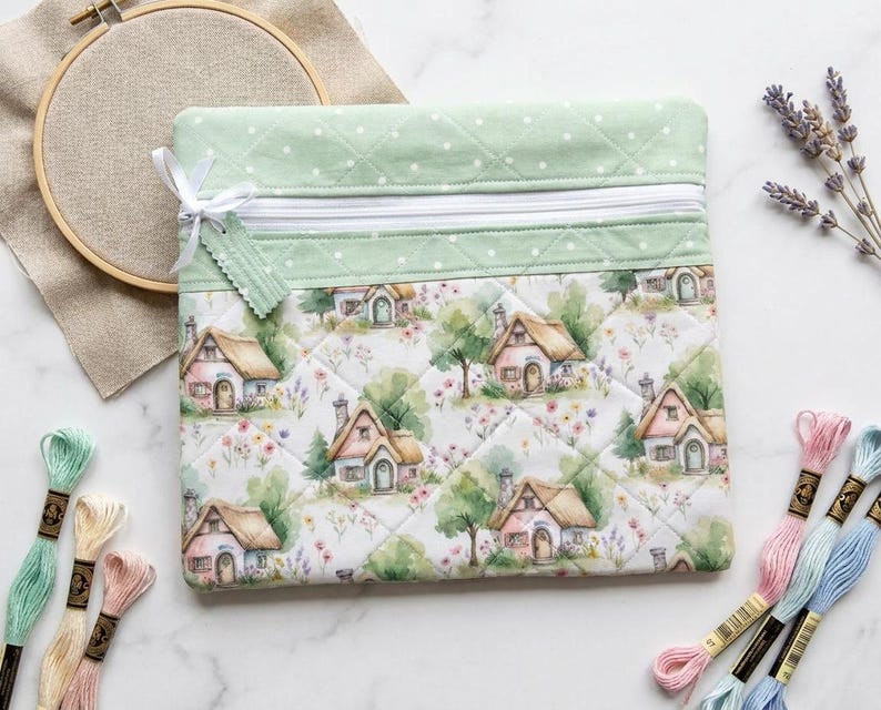 May include: A square quilted zipper pouch featuring a light green fabric top with white polka dots and a main body printed with a pattern of small cottages, trees, and flowers. The pouch includes a white zipper closure with a small fabric tag.