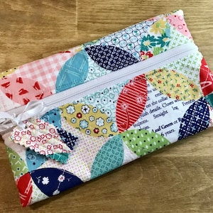May include: A colorful, quilted pencil case with a white zipper and a small fabric tag tied with a white ribbon. The case features a patchwork design with various floral and geometric patterns in red, blue, yellow, and green. The case is approximately 9 inches long.