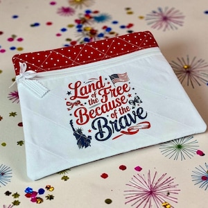 May include: White zippered pouch with a red polka dot top and the text "Land of the Free Because of the Brave" in red, white, and blue. Features an American flag and the Statue of Liberty.