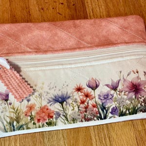May include: A quilted pouch with a floral design. The top is a solid peach color, and the bottom features a white background with a watercolor-style flower garden. A small tag is attached with a white ribbon.