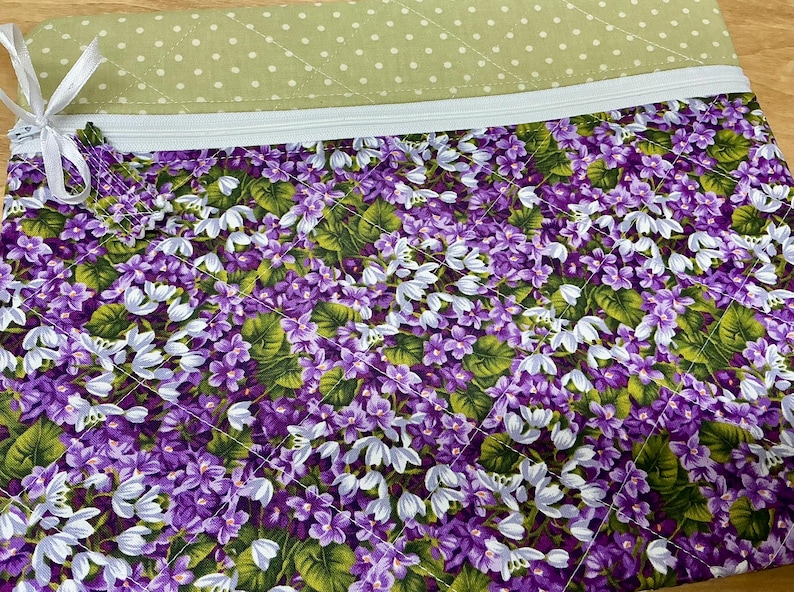 Puede incluir: Una bolsa con cremallera de estampado floral morado y blanco con un forro de tela verde con lunares. La bolsa est&aacute; hecha de tela acolchada.