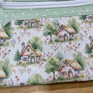 May include: A quilted zipper pouch featuring a repeating watercolor print of small cottages with thatched roofs, surrounded by green trees and colorful wildflowers. The top border is a light green fabric with white polka dots and a white zipper.