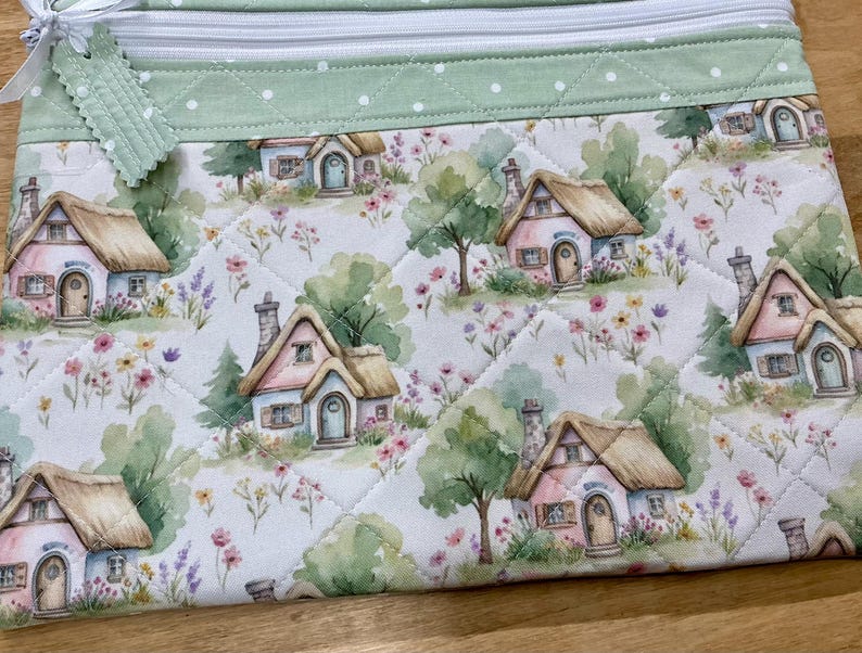 May include: A quilted zipper pouch featuring a repeating watercolor print of small cottages with thatched roofs, surrounded by green trees and colorful wildflowers. The top border is a light green fabric with white polka dots and a white zipper.