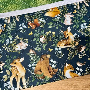 May include: A quilted fabric pouch with a woodland animal print. The design features deer, rabbits, foxes, squirrels, and birds amidst foliage and flowers on a dark blue background. The pouch has a white zipper and a green patterned lining.