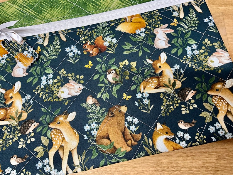 May include: A quilted fabric pouch with a woodland animal print. The design features deer, rabbits, foxes, squirrels, and birds amidst foliage and flowers on a dark blue background. The pouch has a white zipper and a green patterned lining.