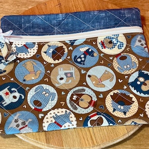 May include: A quilted fabric pouch with a zipper closure. The pouch features a repeating pattern of dog illustrations in blue and brown circles. The top section is a dark blue denim-like fabric. A white ribbon is attached to the zipper pull.