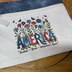 May include: White quilted zippered pouch with a blue denim top. The pouch features a floral design with the word "AMERICA" in colorful letters, surrounded by flowers and butterflies. The words "Freedom, Brave, Independence, Power" are also included.