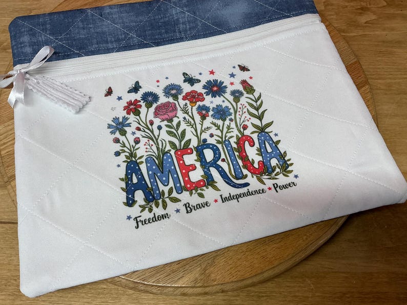 May include: White quilted zippered pouch with a blue denim top. The pouch features a floral design with the word "AMERICA" in colorful letters, surrounded by flowers and butterflies. The words "Freedom, Brave, Independence, Power" are also included.