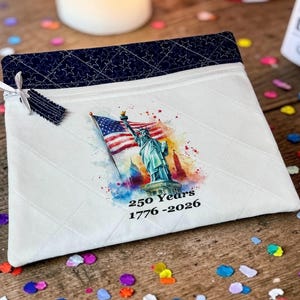 May include: A white quilted pillowcase with a navy blue star-patterned border. The pillowcase features a watercolor image of the Statue of Liberty and an American flag. Text on the pillowcase reads "250 Years 1776-2026".