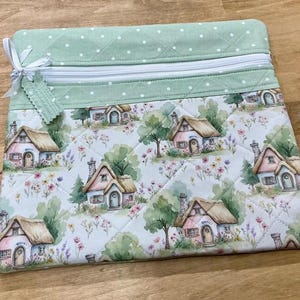 May include: A quilted zippered pouch featuring a light green top panel with white polka dots and a main body panel displaying a repeating watercolor pattern of small cottages with thatched roofs, trees, and colorful wildflowers, finished with a white ribbon.