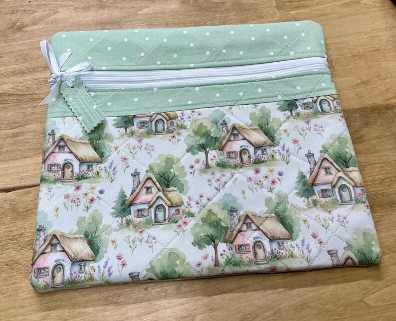 May include: A quilted zippered pouch featuring a light green top panel with white polka dots and a main body panel displaying a repeating watercolor pattern of small cottages with thatched roofs, trees, and colorful wildflowers, finished with a white ribbon.