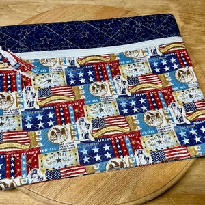 Pu&ograve; includere: Una pochette rettangolare in tessuto trapuntato con chiusura a cerniera. La pochette presenta un design patriottico con bandiere americane, stelle e la scritta "LIBERTY AND JUSTICE FOR ALL". La sezione superiore &egrave; blu navy con stelle bianche.