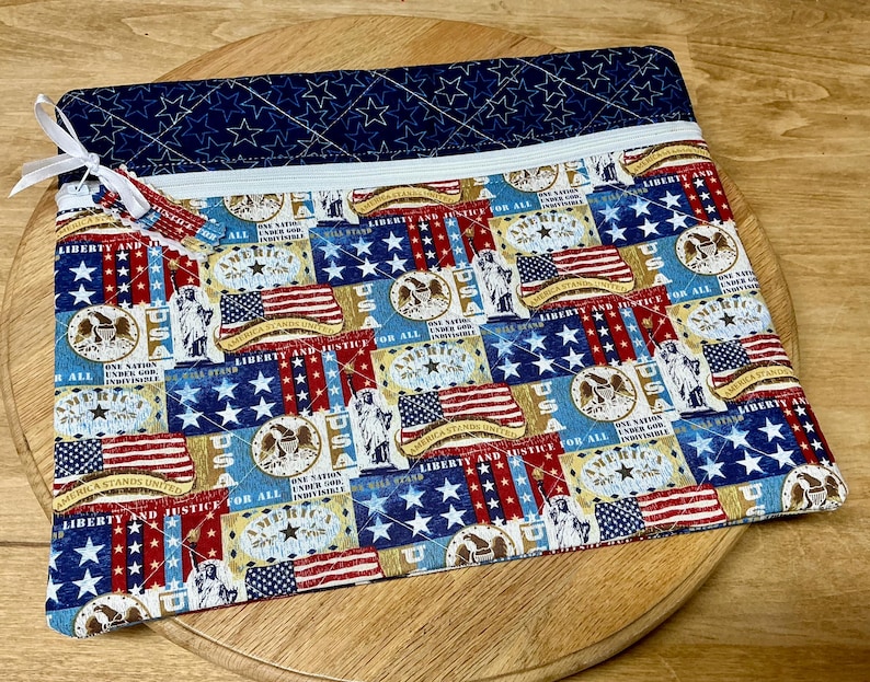 Pu&ograve; includere: Una pochette rettangolare in tessuto trapuntato con chiusura a cerniera. La pochette presenta un design patriottico con bandiere americane, stelle e la scritta "LIBERTY AND JUSTICE FOR ALL". La sezione superiore &egrave; blu navy con stelle bianche.