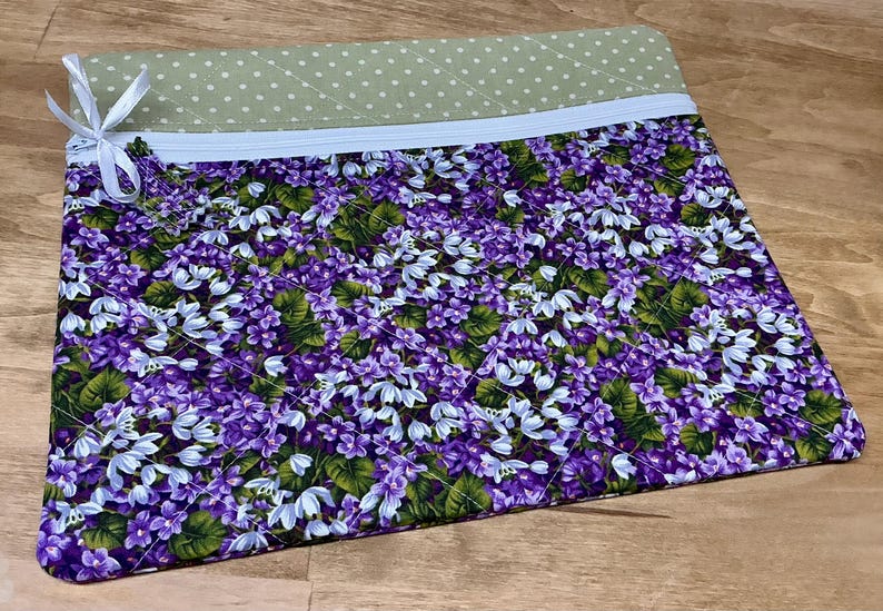 Puede incluir: Una bolsa con cremallera de color morado y verde con estampado floral. La bolsa est&aacute; acolchada y tiene un forro de lunares verde claro.