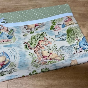 May include: A quilted pillowcase featuring a light blue zipper and a light green polka dot border. The main fabric displays a colorful cartoon scene with Winnie the Pooh, Tigger, and Eeyore. A light blue ribbon is tied to the side.