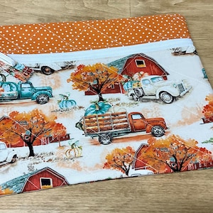 May include: A white zippered pouch with a quilted orange and white polka dot fabric lining. The front of the pouch features a white background with a repeating pattern of red barns, fall trees, and vintage trucks carrying pumpkins.