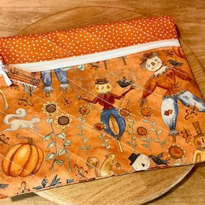 May include: Orange quilted fabric pouch with a zipper closure and white polka dot trim. The pouch features a fall-themed print with scarecrows, sunflowers, pumpkins, and squirrels. A white ribbon is attached to the zipper pull.
