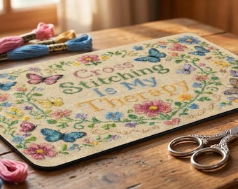 Puede incluir: Una alfombrilla rectangular con el texto Cross Stitching Is My Therapy rodeado de un colorido patrón de flores y mariposas. La alfombrilla se acompaña de varias madejas de hilo de bordar, unas tijeras plateadas y un dedal de metal.