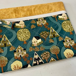 May include: A quilted fabric pouch with a teal background featuring a repeating pattern of houses and trees in shades of green, brown, and cream. The top edge is a mustard yellow fabric with a white zipper and a small white ribbon. The pouch is approximately 10 inches wide and 8 inches tall.