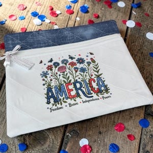 May include: A white quilted pouch with a denim blue top edge. The pouch features the word "AMERICA" in red, white, and blue, surrounded by floral and butterfly illustrations. The words "Freedom, Brave, Independence, Power" are below.