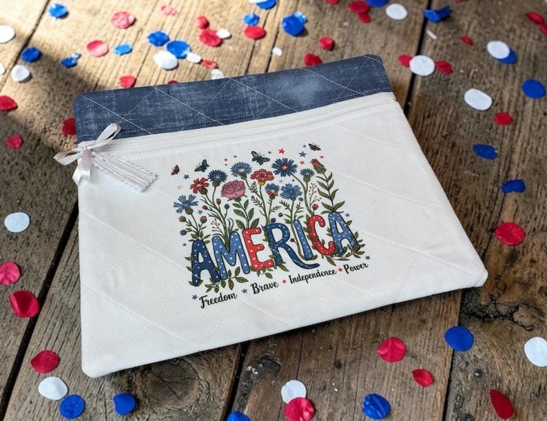 May include: A white quilted pouch with a denim blue top edge. The pouch features the word "AMERICA" in red, white, and blue, surrounded by floral and butterfly illustrations. The words "Freedom, Brave, Independence, Power" are below.