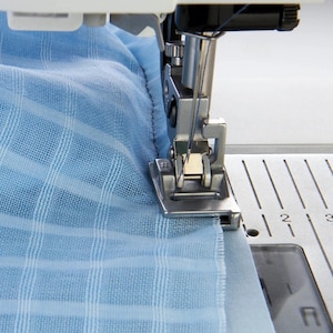 Puede incluir: Primer plano de una máquina de coser en acción, cosiendo tela azul claro. La aguja y el prensatelas son visibles, creando una costura en el material a cuadros. La máquina está sobre una superficie blanca.