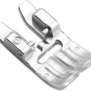 May include: Silver sewing machine foot with a clear plastic guide. The foot has a small hole on the side and a larger opening in the centre.