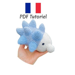 Puede incluir: Un juguete amigurumi de crochet azul y blanco con forma de criatura espinosa. El juguete está siendo sostenido en una mano. El texto "PDF Tutorial" es visible sobre el juguete.
