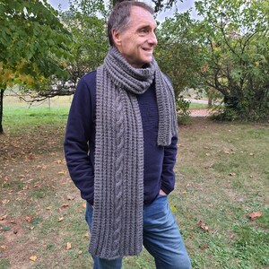 Può includere: Una sciarpa di lana grigia con un motivo a treccia. La sciarpa è indossata da una persona che indossa un maglione blu scuro e jeans blu.