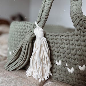 Puede incluir: Una bolsa de mano de crochet de color verde salvia con dos borlas blancas y verde salvia. La bolsa tiene un asa trenzada y un diseño tejido.