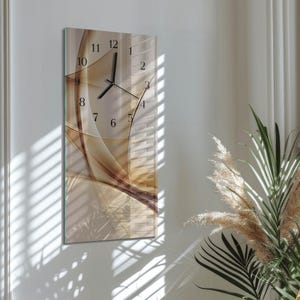 Può includere: Un orologio da parete rettangolare con un design astratto beige e marrone. Il quadrante dell'orologio presenta numeri e lancette nere. L'orologio è montato su una parete bianca, con luce naturale e piante sullo sfondo.