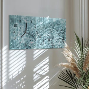 May include: A rectangular wall clock with a teal and white nature-themed design. The clock face features black numerals and hands, indicating the time. The background shows a blurred image of grass with water droplets.