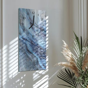 May include: A rectangular wall clock with a blue and gray marble design. The clock face features black numerals and hands. The clock is mounted on a white wall, with natural light and plants in the background.
